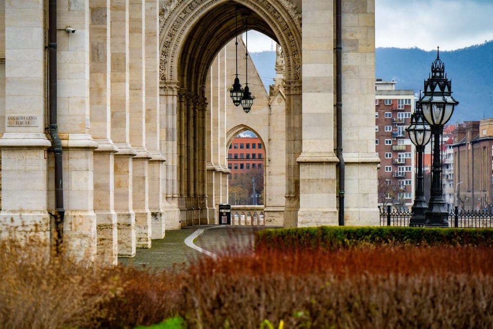 A view through a grand stone archway in Budapest, Hungary, showing a pathway, street lamps, and distant buildings and mountains.