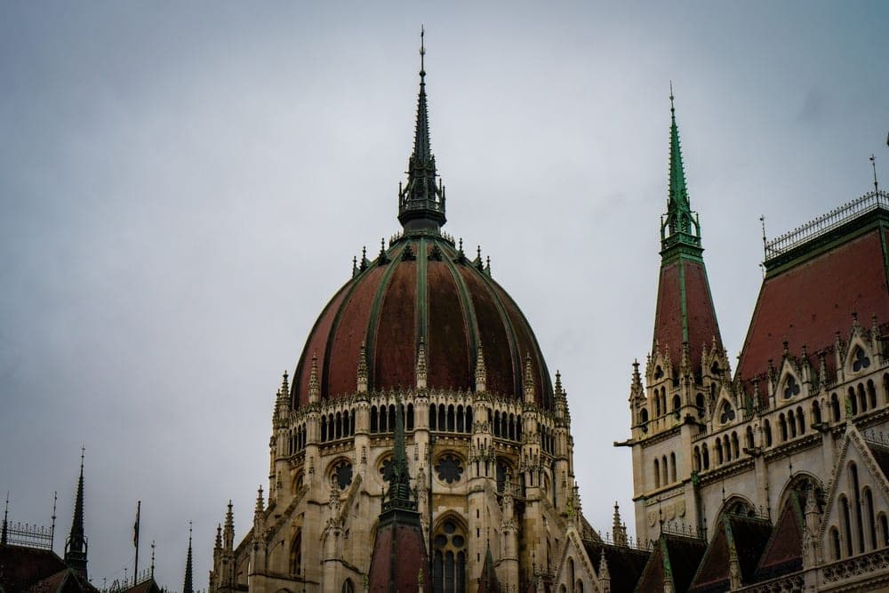 The Hungarian Parliament Building in Budapest, Hungary, with its large copper dome and a tall green spire against a cloudy sky.