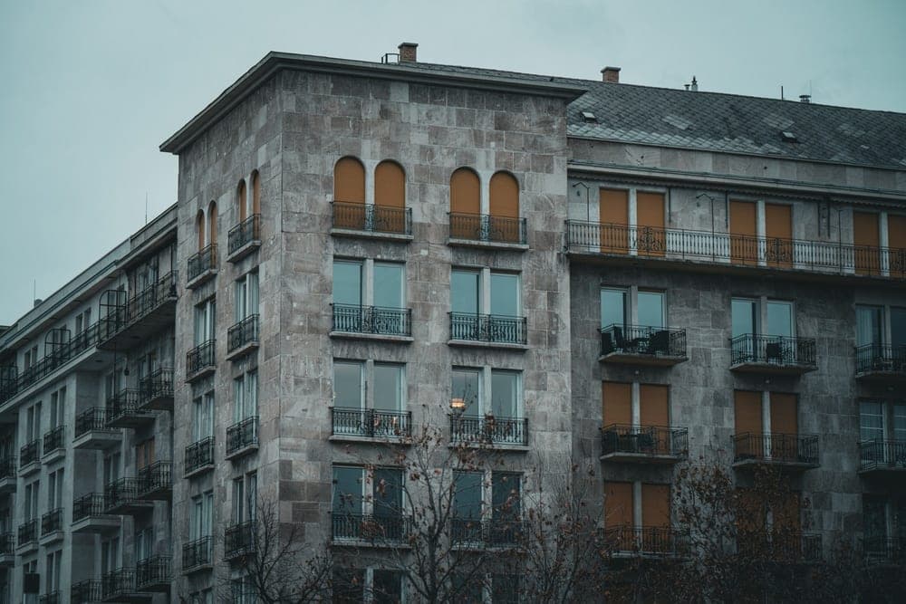 A grey stone apartment building in Budapest, Hungary, with arched windows, wrought-iron balconies, and burnt orange shutters, under an overcast sky.