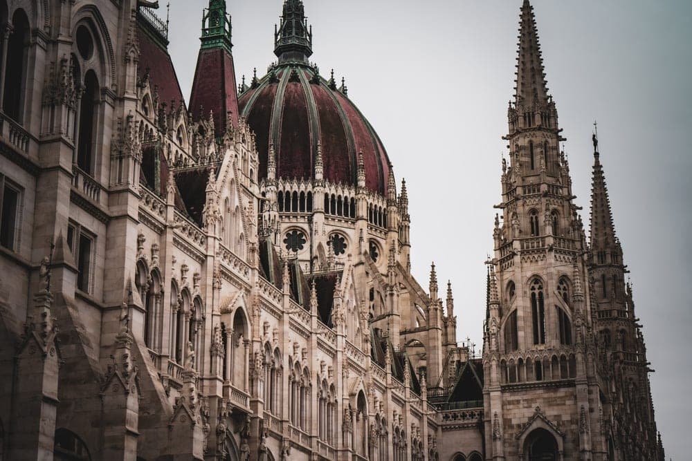 The Hungarian Parliament Building in Budapest, Hungary, showcasing its Gothic Revival architecture with a prominent red dome and spires under an overcast sky.