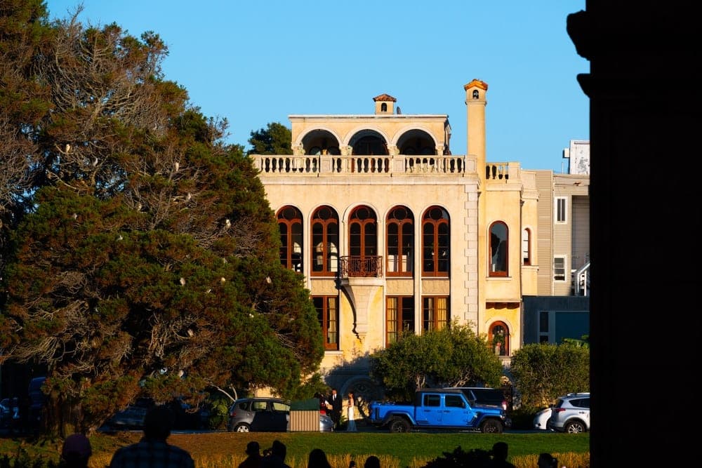 A golden mansion with arched windows and a large evergreen tree in front, on a sunny day.