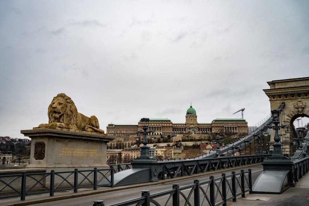 The Széchenyi Chain Bridge in Budapest, Hungary, with a stone lion statue in the foreground and Buda Castle in the background under an overcast sky.