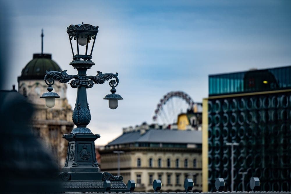 An ornate dark teal lamppost stands in the foreground with the Budapest skyline, including a Ferris wheel and grand architecture, in the blurred background.