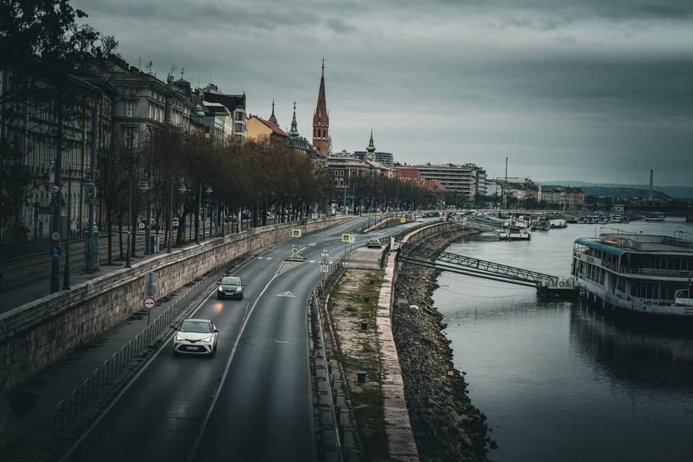 The Danube River embankment in Budapest, Hungary, with cars on the road, historic buildings, and docked boats under a cloudy sky.