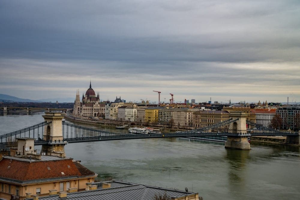 The Chain Bridge and Parliament Building over the Danube River in Budapest, Hungary, under a cloudy sky.