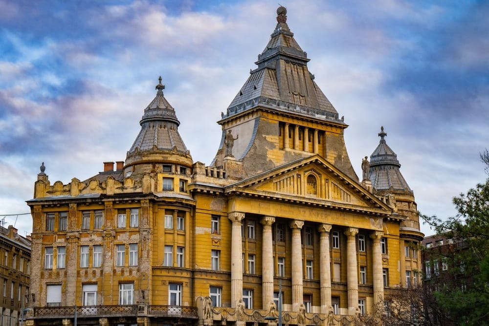 The ornate, yellow facade of the Hungarian National Bank building in Budapest, Hungary, under a dramatic cloudy sky.