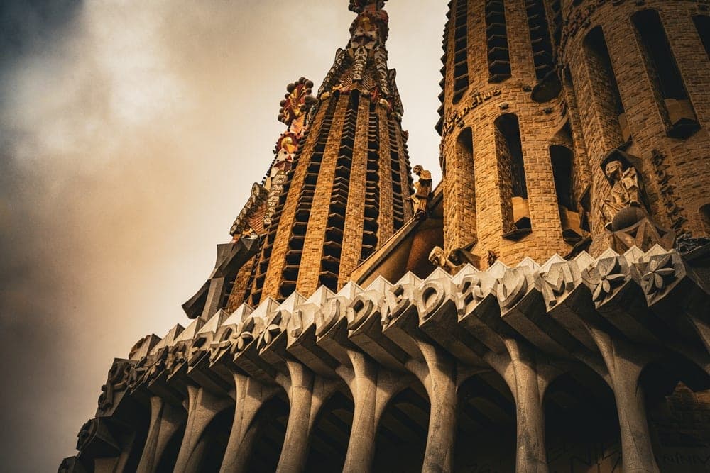 The Sagrada Familia basilica in Barcelona, Spain, viewed from a low angle, showcasing its intricate spires and stone carvings against a dramatic sky.