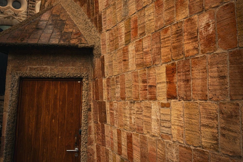 A close-up of a textured brick wall and a wooden door, with the Sagrada Família in Barcelona, Spain, in the background.