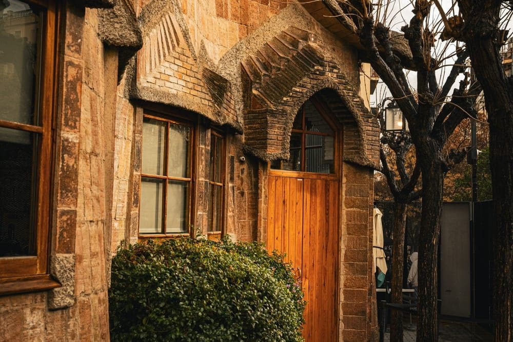 Exterior of a brick building with a wooden door and arched window, flanked by bare trees in Barcelona, Spain.