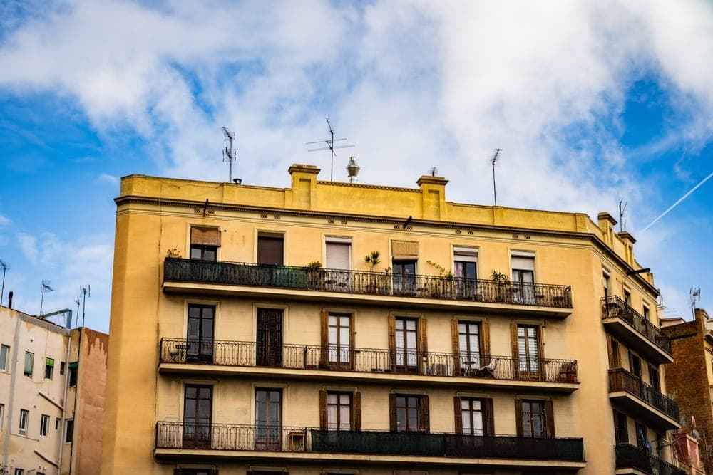 Ochre-yellow apartment building facade with multiple balconies and windows under a bright, cloudy blue sky in Barcelona, Spain.