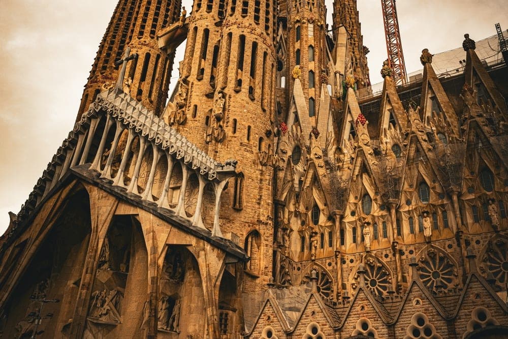 The Sagrada Familia basilica in Barcelona, Spain, seen from a low angle against a dramatic, cloudy sky, with a construction crane in the background.