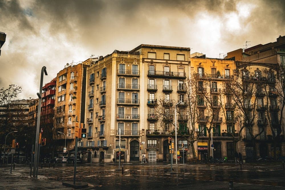 Apartment buildings with ornate balconies on a wet street in Barcelona, Spain, under a cloudy sky.