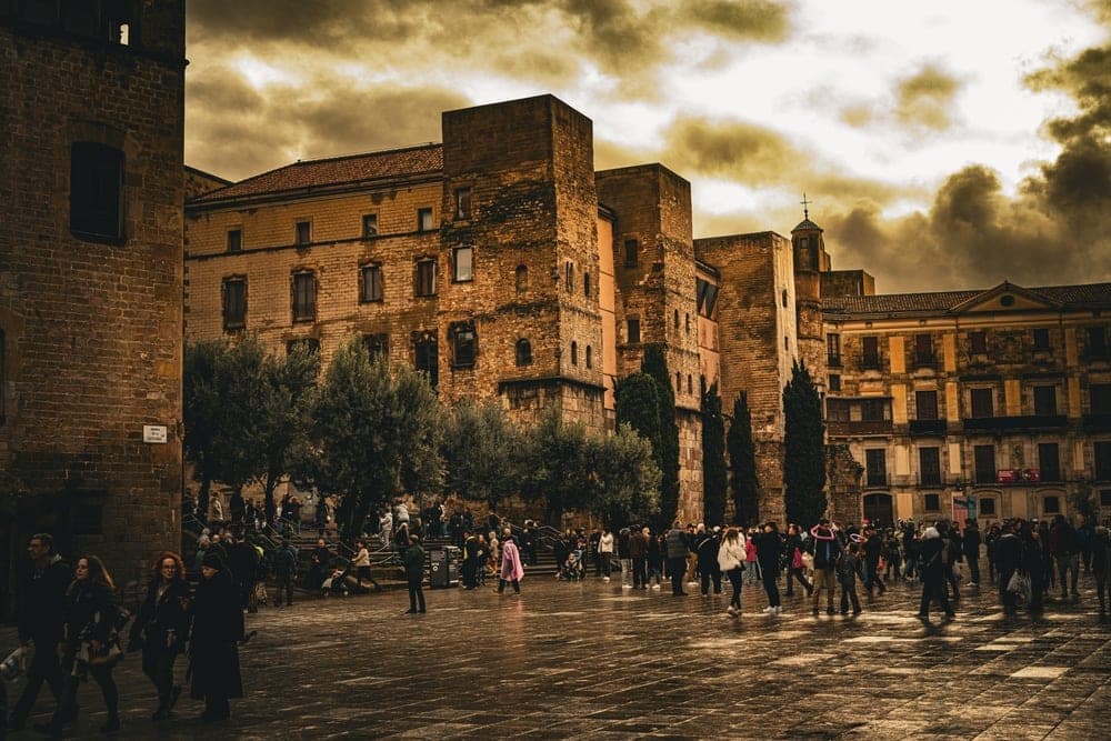 People walking in a wet cobblestone plaza in front of historic stone buildings in Barcelona, Spain, under a dramatic cloudy sky.