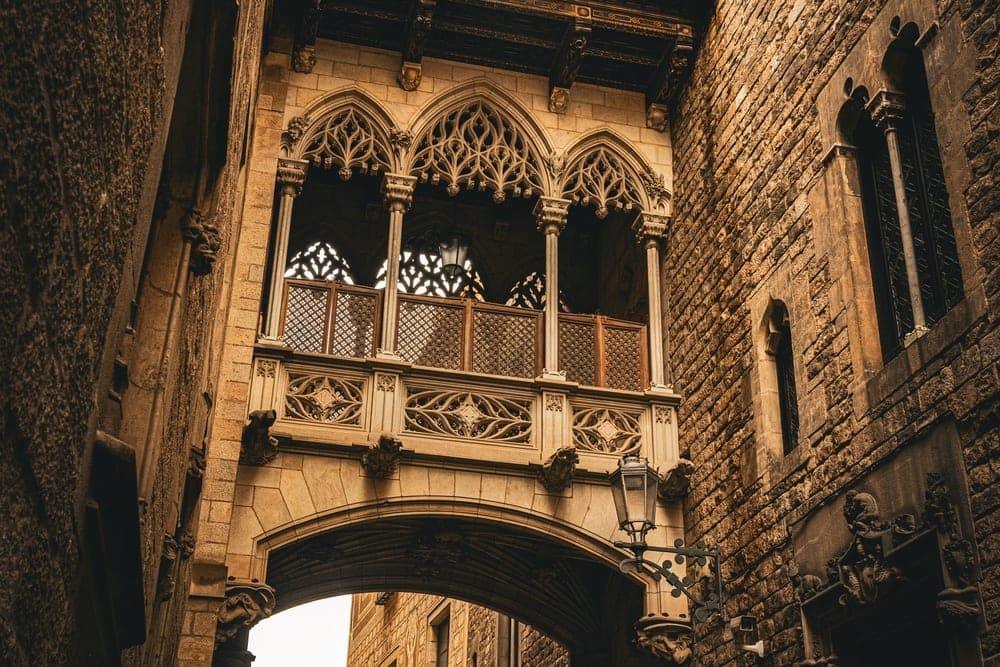 The Pont del Bisbe, a stone Gothic bridge connecting two buildings over a street in Barcelona, Spain, with people walking below.