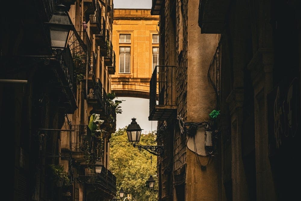 A narrow, wet street in Barcelona, Spain, with tall buildings on either side and a yellow archway overhead.