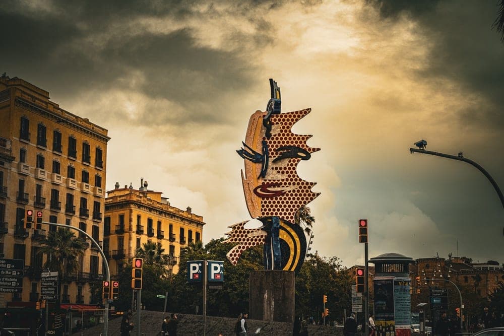 The 'Barcelona Head' sculpture by Roy Lichtenstein, a colourful pop-art face, stands in front of yellow buildings under a dramatic cloudy sky in Barcelona, Spain.