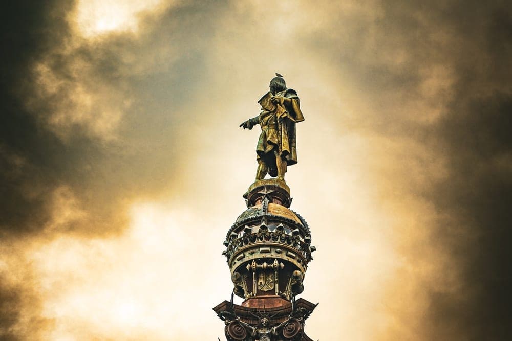 The Columbus Monument in Barcelona, Spain, with a statue of Christopher Columbus pointing seaward, set against a dramatic, dark, and cloudy sky with golden light.