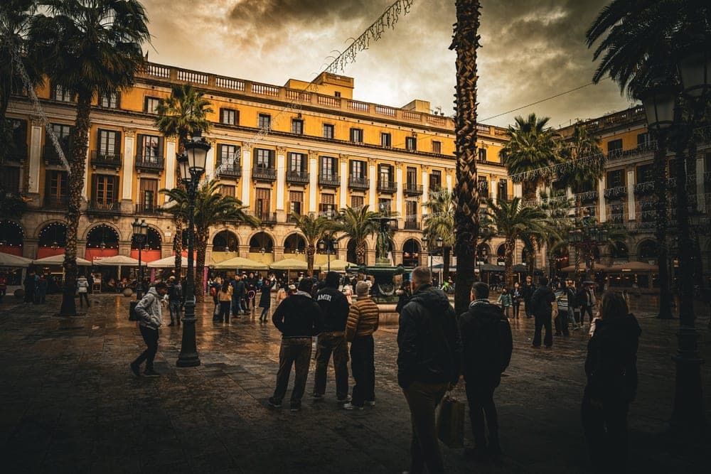 People gathered in Plaça Reial, Barcelona, Spain, under a dramatic, cloudy sky and with ornate yellow buildings in the background.