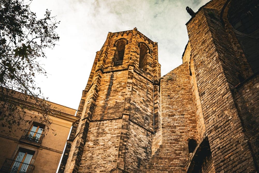 The historic stone tower of the Cathedral of Barcelona, Spain, viewed from below against a dramatic cloudy sky.