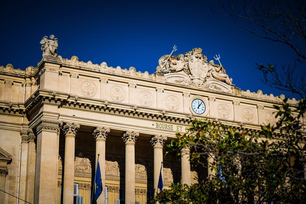 The Beaux-Arts facade of the Palais de la Bourse in Marseille, France, with its columns, sculptures, and clock, under a clear blue sky.