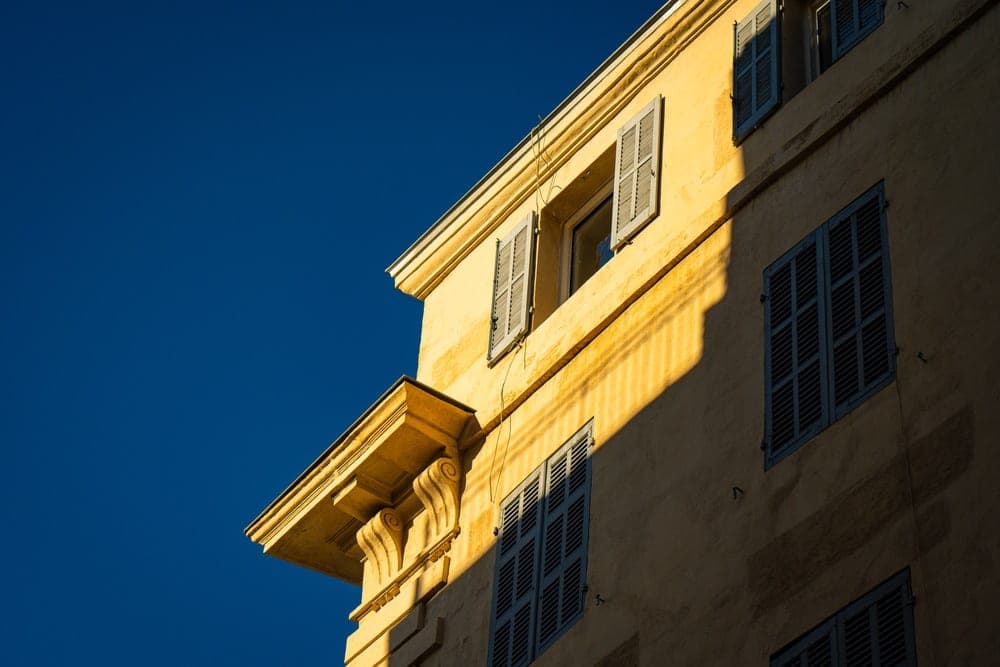Building facade with blue shutters and dramatic shadows under a clear blue sky in Marseille, France.