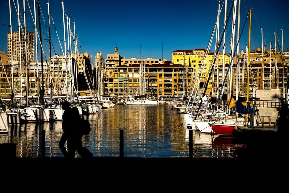 Silhouettes of sailboats and a walker on the Quai De Rive-Neuve in Marseille, France, with colourful buildings reflected in the water.
