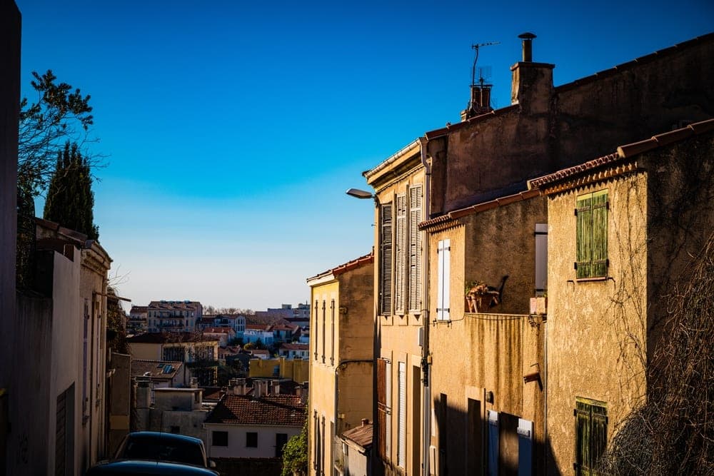A view down a sunlit street in Marseille, France, with traditional stucco buildings and terracotta roofs under a bright blue sky.