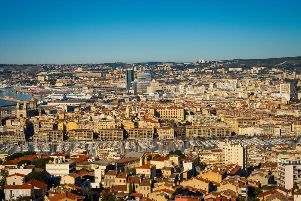 Panoramic view of Marseille, France, showing the city's dense urban landscape, harbour with yachts, and distant hills under a clear blue sky.