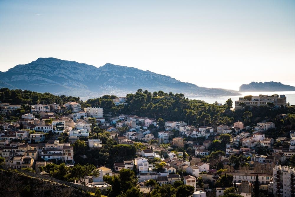 Panoramic view of the residential hillsides of Marseille, France, with numerous white buildings and terracotta roofs cascading down a slope, backed by a vast mountain range and the blue sea under a clear sky.