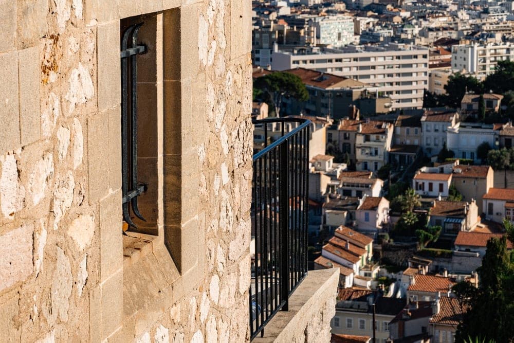 View from a stone building's balcony overlooking the city of Marseille, France, with mountains in the background under a clear blue sky.