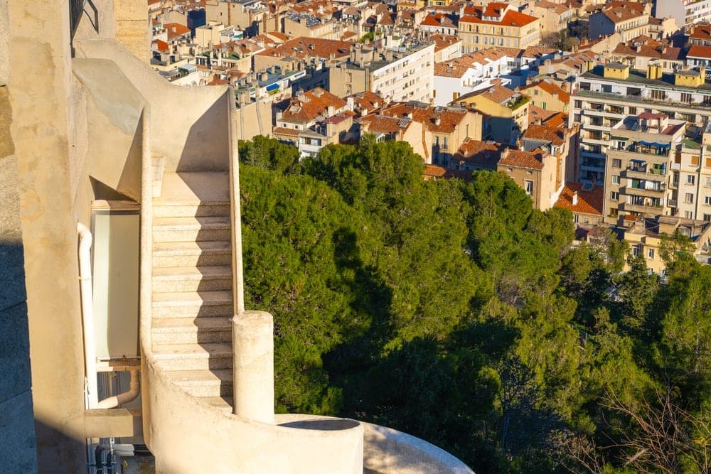 A sunlit concrete spiral staircase in Marseille, France, overlooking a dense cityscape.