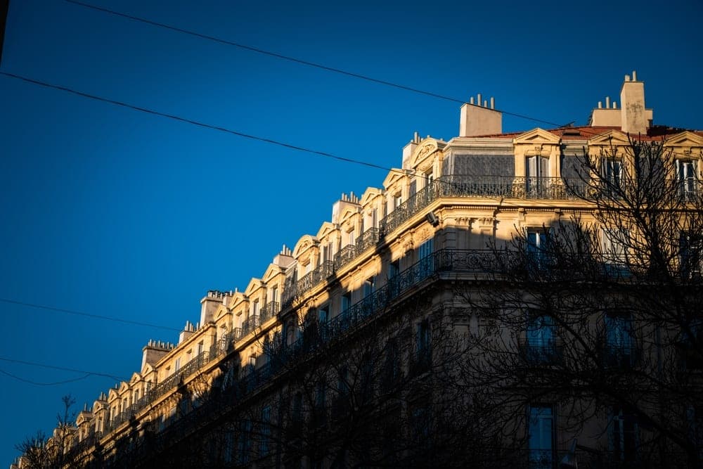 A sunlit Haussmannian building in Marseille, France, with ornate balconies and a silhouetted tree in the foreground against a blue sky.