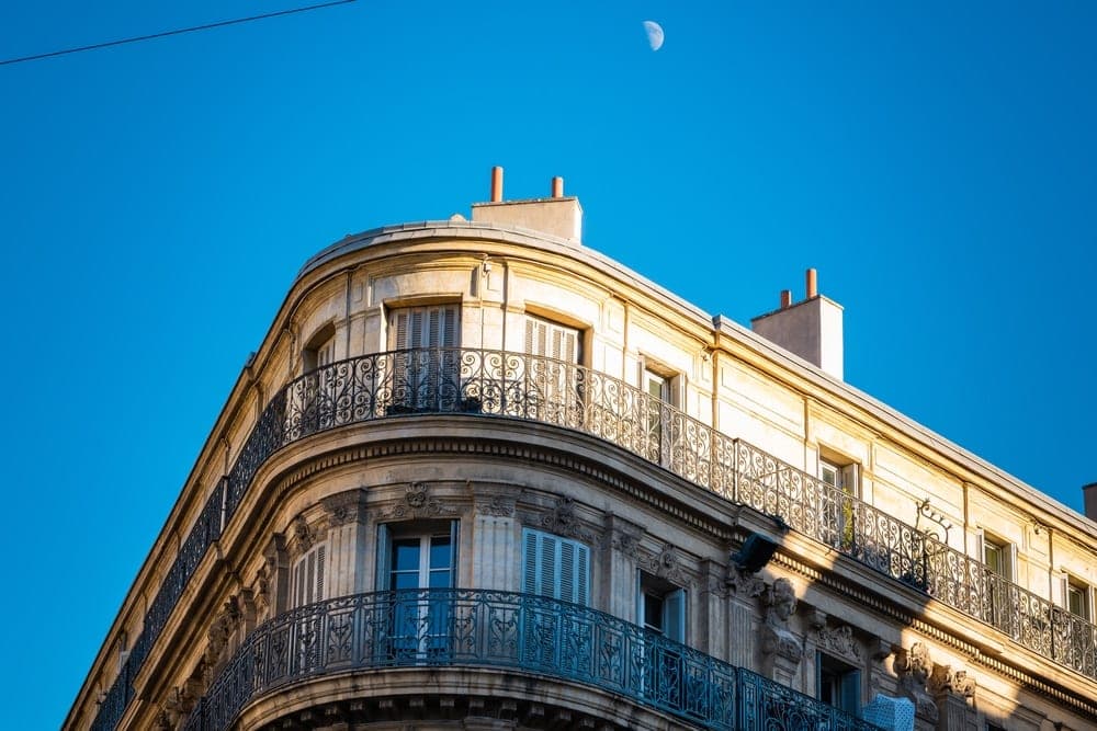 Ornate building corner with blue shutters and balconies in Marseille, France, under a clear blue sky with a crescent moon.