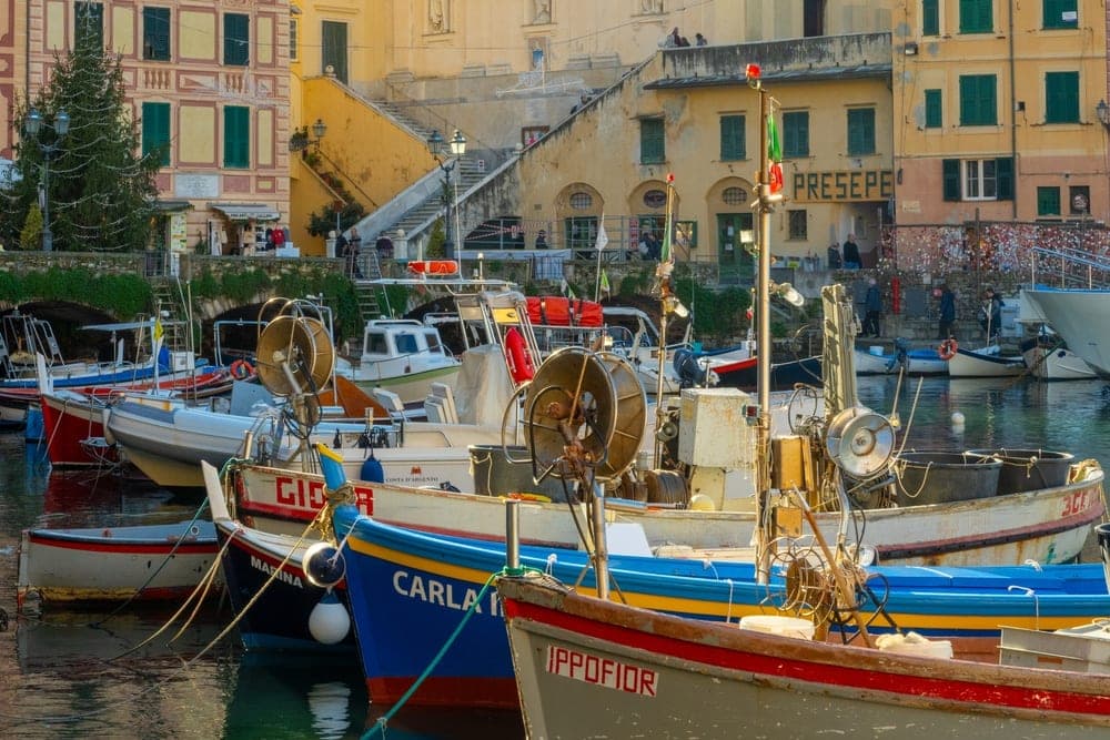 Fishing boats moored in the harbour of Camogli, Genoa, Italy, with colourful buildings and a church in the background under a clear blue sky.