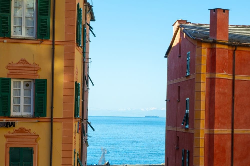 View down a narrow street in Portofino, Italy, flanked by colorful buildings, leading to a harbor with boats and the blue sea.