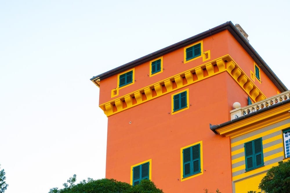 A bright orange building with yellow trim and green shutters, partially obscured by lush green trees, in Portofino, Italy.