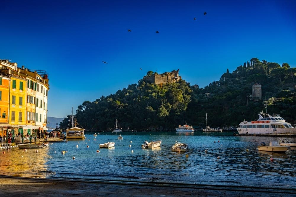 The picturesque harbour of Portofino, Italy, with colourful buildings on the left, boats on the water, and Castello Brown on a green hillside under a bright blue sky.
