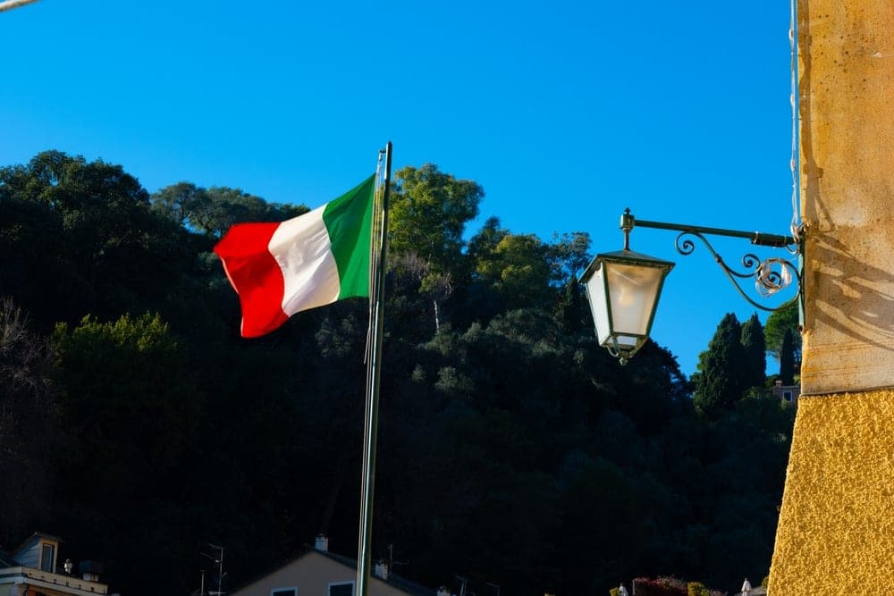 The Italian flag waves against a bright blue sky, with a vintage lantern and a green hillside in Portofino, Italy.