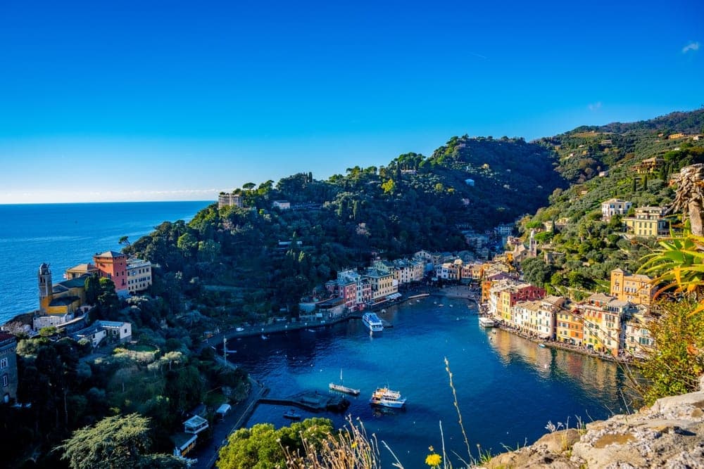 A scenic view of Portofino, Italy, with colorful buildings lining a blue harbor and lush green hillsides.