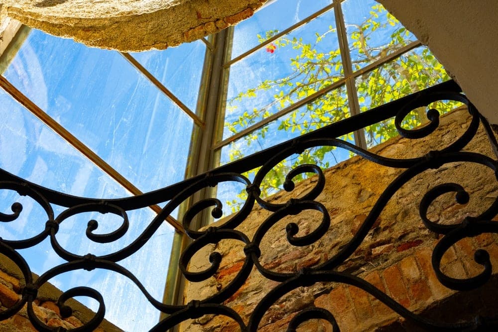 Wrought-iron railing in front of a sunlit stone wall and a window with blue sky and green foliage, Portofino, Italy.