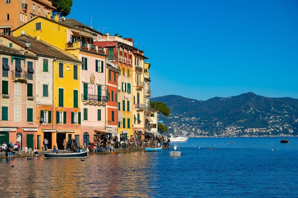 Colorful buildings lining the waterfront of Portofino, Italy, with boats in the blue harbor and hills in the background.