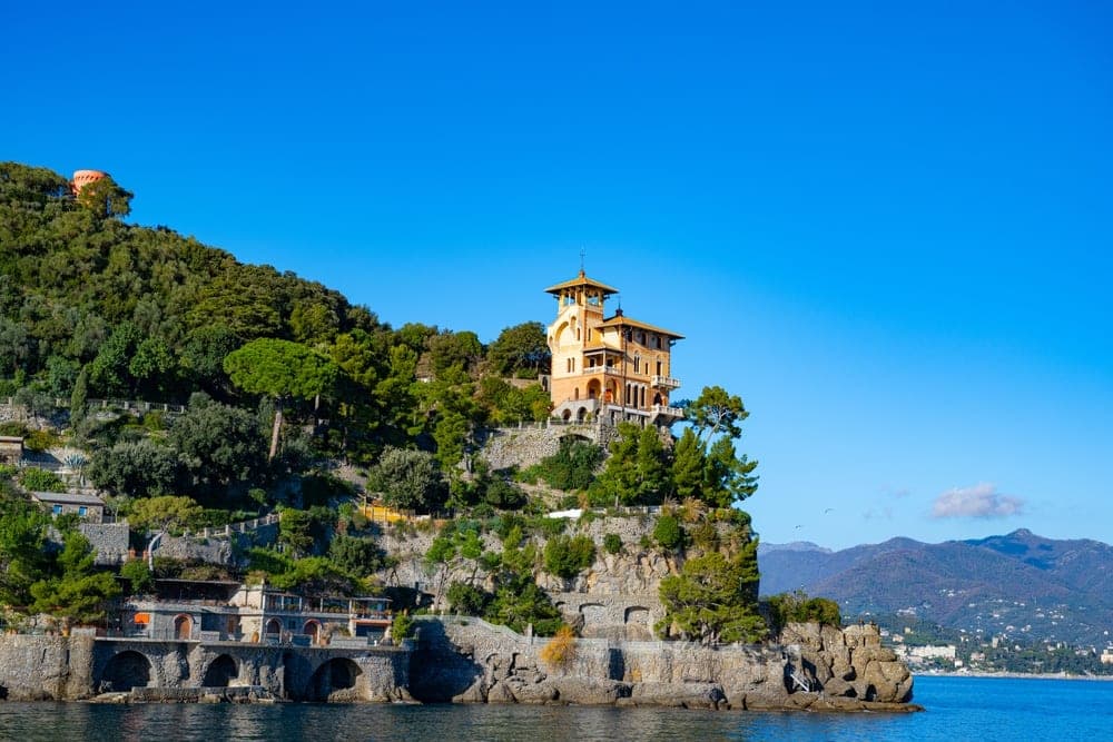 A terracotta villa sits atop a lush green cliff overlooking the blue sea in Portofino, Italy.