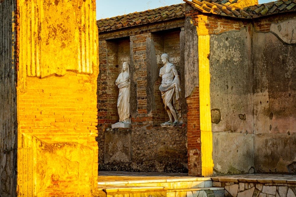 Two ancient marble statues, a male and a female, stand in niches within a brick wall at Pompeii, Italy, illuminated by golden afternoon light.