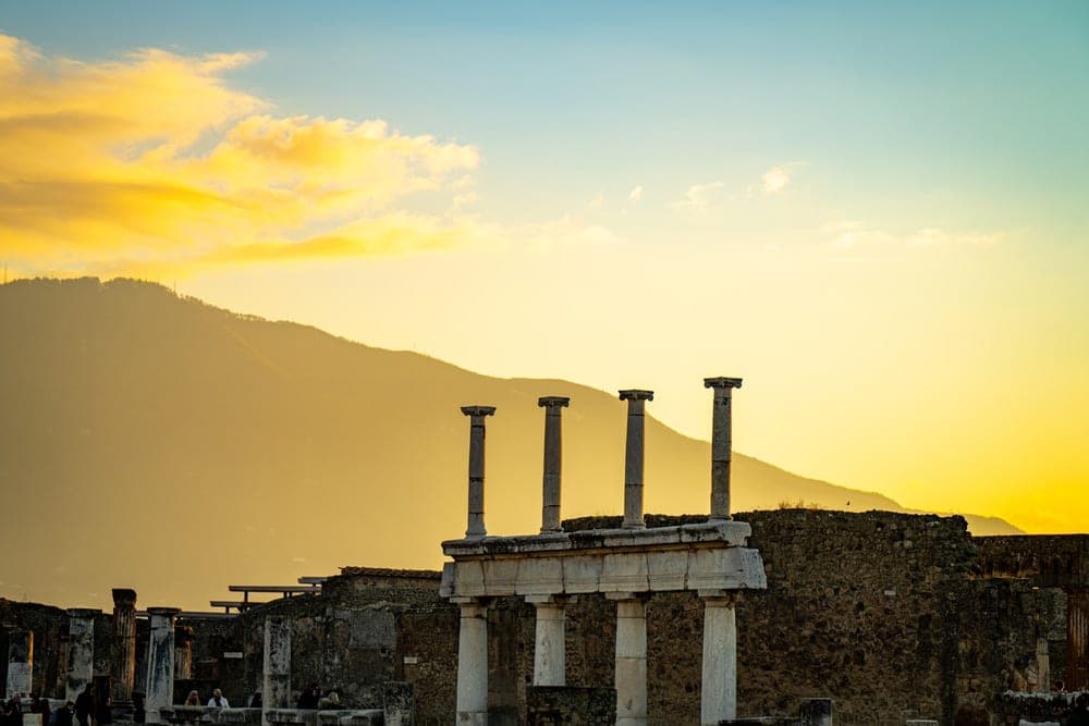 Ancient Roman columns in Pompeii, Italy, silhouetted against a vibrant sunset sky with Mount Vesuvius in the background.