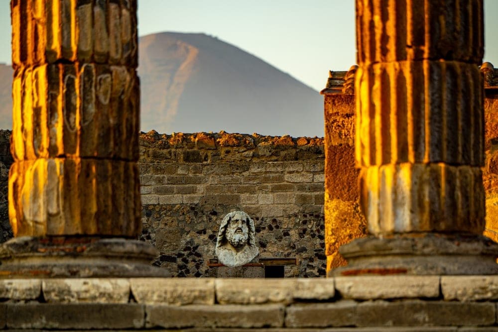 Bust of a bearded man set against a stone wall in Pompeii, Italy, with ancient columns in the foreground and Mount Vesuvius in the background.