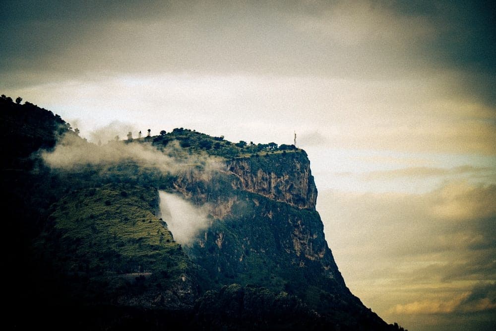 A steep, rugged cliff face covered in green vegetation and shrouded in mist, in Palermo, Italy.