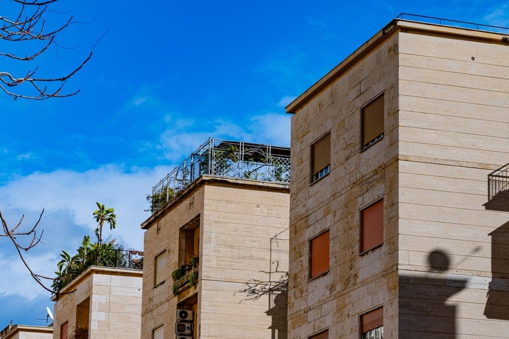 Modern apartment buildings with travertine facades and rooftop gardens under a bright blue sky in Palermo, Italy.