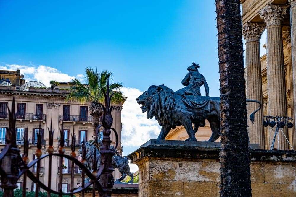 Bronze sculpture of a lion with a rider in Palermo, Italy, with classical architecture and palm trees in the background.