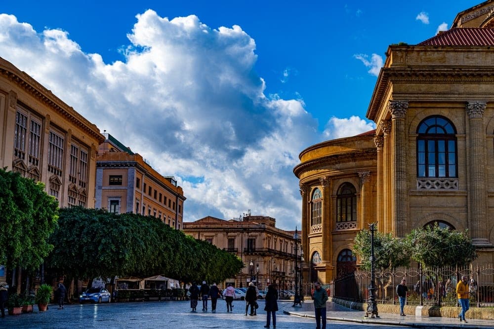A view of a sunlit piazza in Palermo, Italy, with ornate ochre-coloured buildings and lush green trees under a dramatic blue sky with white clouds.