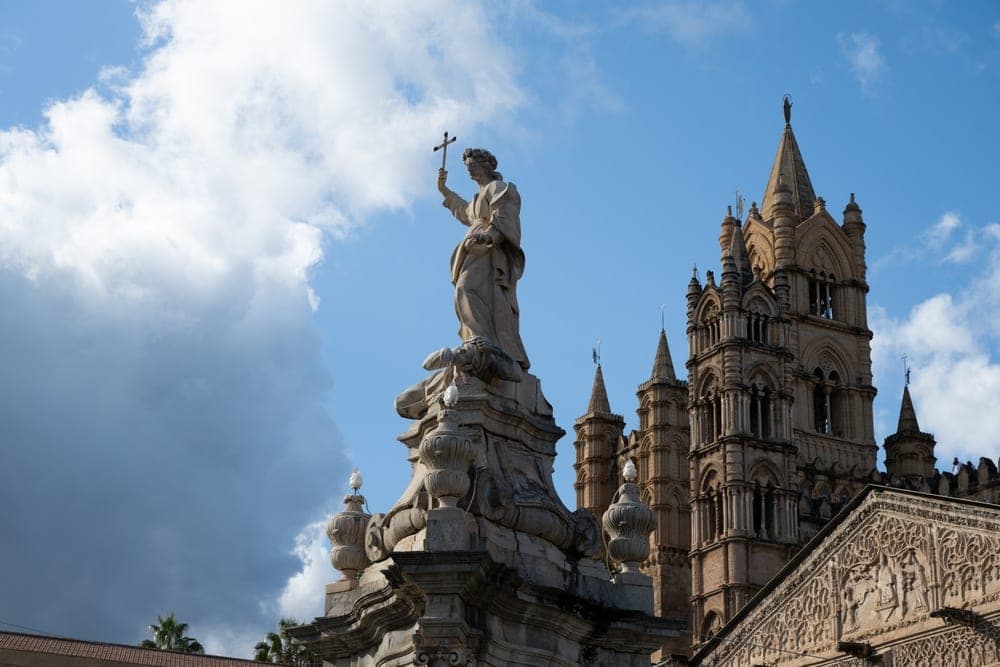 A statue holding a cross stands before the Palermo Cathedral in Palermo, Italy, under a dramatic sky.